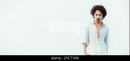 Young asian man with curly hair wearing a light, unbuttoned linen shirt stands confidently against a clean, white background. Ample copy space area to Stock Photo