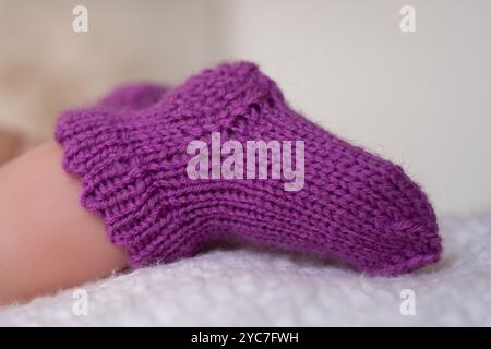 A pair of cozy purple knitted booties for infants is placed delicately on a soft white blanket, showcasing their adorable texture and warmth. Stock Photo