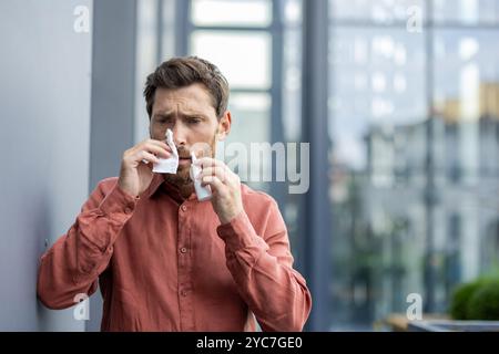 Man using tissue outdoors in urban setting, wearing casual shirt, looking concerned. Scene depicts health issues like cold or allergies, reflecting ev Stock Photo