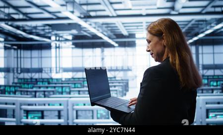 Computer scientist doing yearly maintenance using laptop to prolong data center equipment life span. IT specialist in server hub facility monitoring server infrastructure using device, camera B Stock Photo