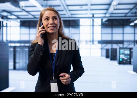 Smiling person overseeing operations in cutting edge data center, coordinating tasks during telephone call. Happy IT specialist in server hub managing systems and communicating via smartphone Stock Photo