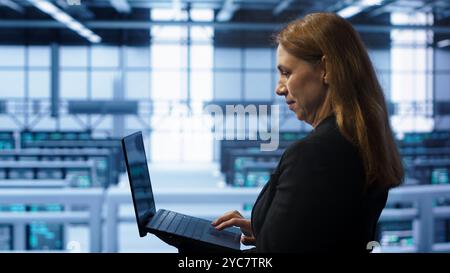 System administrator in server hub checking recovery plan on laptop, monitoring energy consumption. IT expert uses notebook in data center workspace, ensuring rigs are functioning optimally, camera A Stock Photo