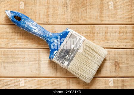 A closeup view of a used paintbrush with blue handle and white bristles lying on a wooden surface. The brush shows signs of wear and tear, with white Stock Photo