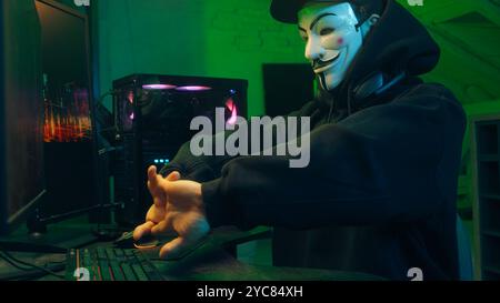 Lviv, Ukraine - June 03, 2024: Masked man anonymous stretching fingers programmer hacking typing computer malware networks web danger scam database Stock Photo
