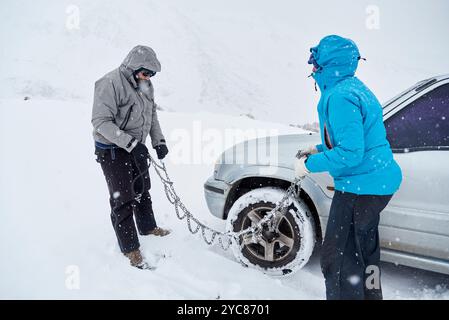 Travel in wintertime, pair of friends, man and woman, putting chains on the wheels of an SUV vehicle, in order to be able to move forward over the sno Stock Photo