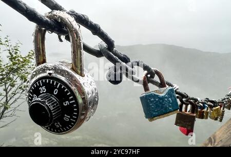 A close-up of multiple padlocks and a combination lock attached to a wet chain outdoors on a rainy day. The misty, natural landscape in the background Stock Photo