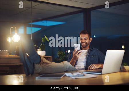 Night, office and man with phone, laptop and online search for social media post at digital agency. Late, relax and creative consultant with Stock Photo