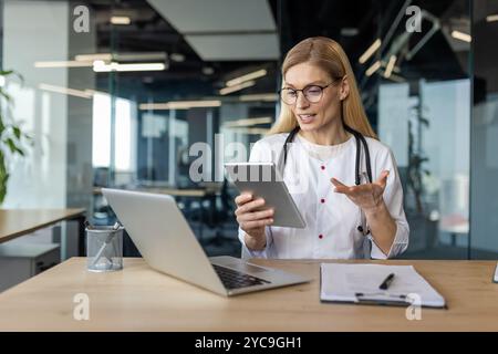 Mature woman doctor uses tablet for video consultation in office. Stethoscope around neck, she interacts confidently, illustrating telemedicine concep Stock Photo