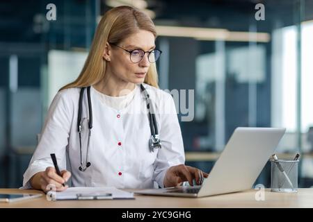 Mature female doctor working on laptop in modern office. Wearing stethoscope, taking notes, conducting research effectively. Emphasizes professionalis Stock Photo