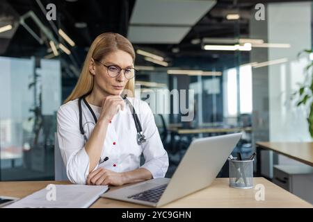 Mature female doctor in office setting using laptop for medical research or patient consultation. Professional attire includes glasses and stethoscope Stock Photo