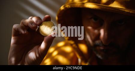 Computer hacker and cryptocurrency scammer in golden yellow hooded jacket holding Bitcoin in hand, selective focus Stock Photo