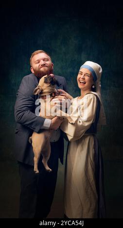 Fun moment of couple, dressed in vintage-style clothing, enjoying playful time with their dog against dark vintage background Stock Photo