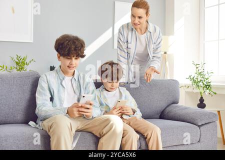 Children brothers sitting on sofa at home holding mobile phones in hands ignoring his angry mother Stock Photo