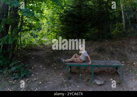 Young boy sits alone on a wooden bench in the woods, surrounded by trees and greenery on a peaceful afternoon.  Stock Photo