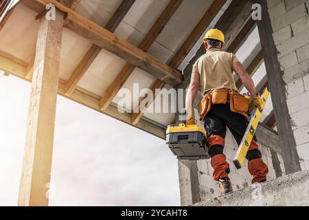 A construction worker stands at a height, holding a toolbox and level, actively assessing a building's framework during the day. Stock Photo