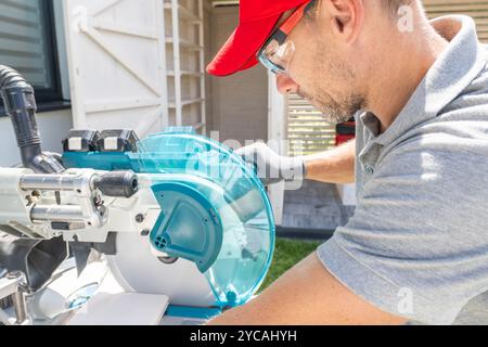 A worker focuses intently on using a miter saw, carefully making precise cuts in wood while wearing protective gear in an outdoor setting. Stock Photo