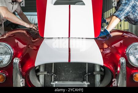 Two individuals are carefully examining the engine compartment of a vintage red sports car in a well-lit garage, focusing on details and conditions. Stock Photo