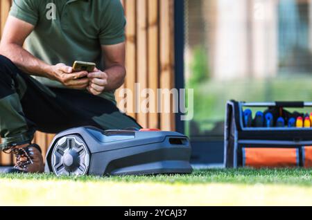 A man kneels beside a robotic lawn mower, using his phone to control it, with a tool set nearby in a contemporary yard. Stock Photo