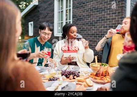 Vibrant scene of group of adult friends enjoying drinks at dinner table outdoors and exchanging presents while celebrating holidays together Stock Photo