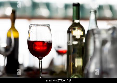 Half-full glass of red wine in a clear stemware goblet surrounded by green and yellow wine bottles. Stock Photo
