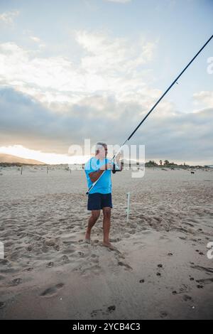 An elderly man engages in fishing on a sandy beach in Valencia, Spain, holding a large fishing rod at dusk with a serene sky in the background Stock Photo