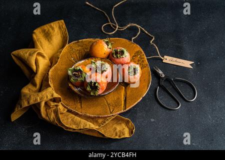 A table decorated in an autumn style with fresh persimmons on a rustic plate, complemented by a linen napkin and scissors, all set against a dark text Stock Photo