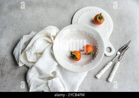 This image features an elegant table setting themed with fresh persimmons, showcasing white decorative plates and a textured gray background Stock Photo