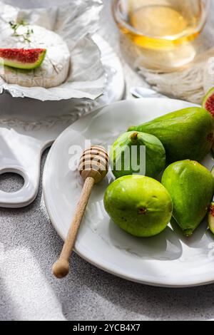 Brie cheese topped with a fig slice, alongside whole green figs and a honey dipper on a white ceramic plate. A glass of white wine complements the sce Stock Photo