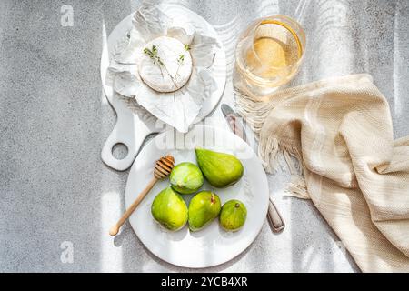 Top view of a gourmet snack with Brie cheese wrapped in paper, paired with ripe green figs and a honey dipper on a white ceramic plate. A glass of whi Stock Photo