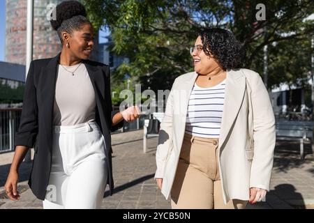 Two professional women enjoying a sunny day while engaging in a friendly conversation. They are both smiling and dressed in business attire, one Afric Stock Photo