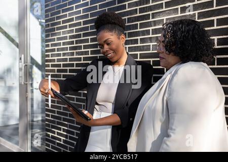 An African American and Hispanic women engage in a discussion while using a tablet outside a modern office building. The younger woman shows the older Stock Photo
