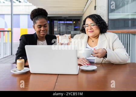 Two women, an African and a Hispanic, are seen collaborating at an outdoor cafe, focused on a laptop screen while one holds a cup of coffee. Stock Photo