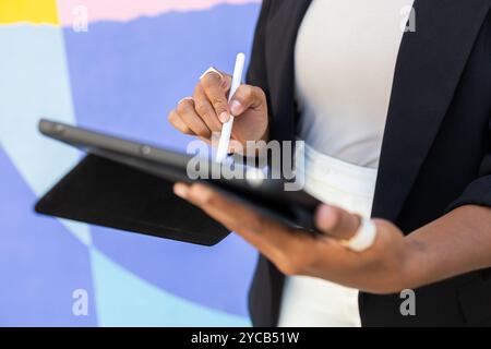 Cropped unrecognizable professional African American woman using a digital tablet and stylus outdoors, focused on the screen against a vibrant multico Stock Photo