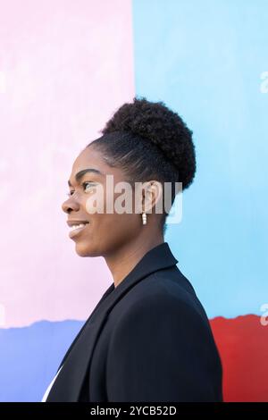 Side view of a smiling African American woman looking away, set against a colorful blue and pink background. She appears confident and stylish in her Stock Photo