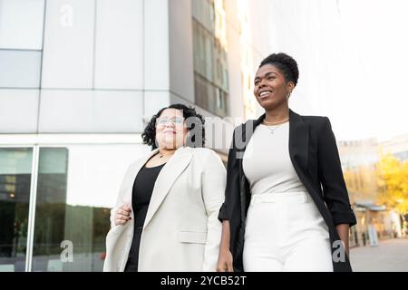 Two professional women, one Hispanic and one African American, walk together through a vibrant urban setting. Engaged in a cheerful conversation, they Stock Photo