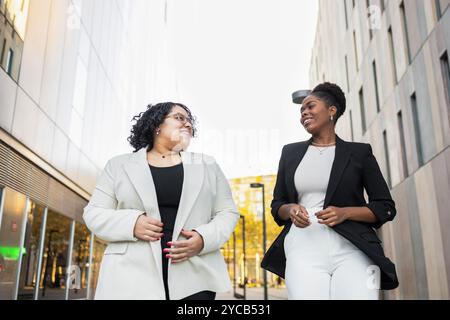 Two professional women, one Hispanic and the other African American, engaging in a cheerful conversation while walking through an urban business distr Stock Photo
