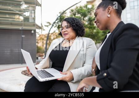 Two businesswomen, one African American and one Hispanic, are engaged in a serious work conversation while reviewing data on a laptop outdoors. Both a Stock Photo