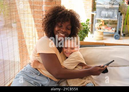 Tender moment between African American mother and her Caucasian son as they enjoy leisure time on a porch, embracing while the son holds a smartphone. Stock Photo