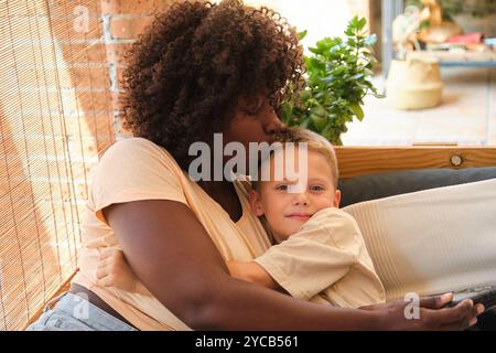 An African American mother kisses her Caucasian son on the forehead while they relax on a couch. Both are looking at each other, in a heartwarming hom Stock Photo