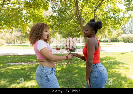 An African American and a Latin American LGBTQIA+ couple share a tender moment in a lush park, exchanging flowers and smiling at each other Stock Photo