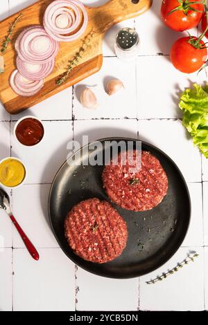 Overhead shot of hamburger patties seasoned with salt and herbs, accompanied by sliced red onions on a wooden cutting board, fresh tomatoes, garlic, l Stock Photo