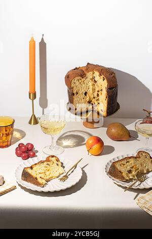 A traditional Italian Panettone displayed on a wooden stand, accompanied by fruits, a candle, and wine, set on a simple, elegantly decorated table wit Stock Photo