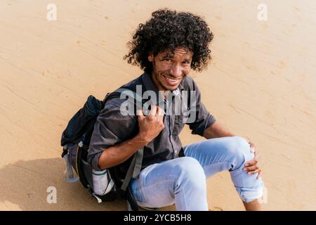 A curly-haired man with a joyful expression, dressed in a casual grey shirt and blue jeans, kneeling on a sandy beach, holds a backpack strap as he lo Stock Photo