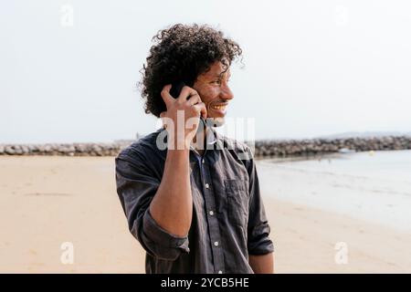 A cheerful man enjoys a conversation on his mobile phone, walking along a tranquil sandy beach, with a calm sea and protective sea wall in the backgro Stock Photo