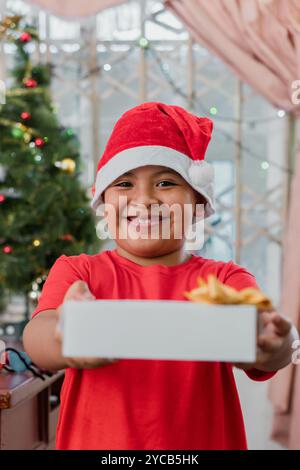Joyful Latin American boy wearing a Santa hat, presenting a gift box with a bow, in front of a decorated Christmas tree, exuding festive cheer Stock Photo