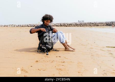 A young man with curly hair sits on a sandy beach, examining the contents of his backpack against a backdrop of calm seas and a distant breakwater Stock Photo