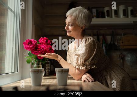 Side view of a mature woman with short gray hair smiles gently while holding and admiring a bouquet of vibrant roses by a window in a rustic kitchen s Stock Photo