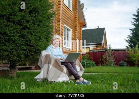 A mature woman sits comfortably in a large garden, working quietly on her laptop next to a log home. She is dressed in a light blue shirt and brown tr Stock Photo