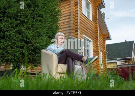 A mature woman engages with a laptop while comfortably seated in a chair outdoors, next to a lush green tree and in front of a log cabin home, looking Stock Photo