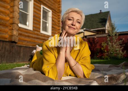 A mature woman in a vibrant yellow jacket found peace amidst loneliness, laying on a blanket in front of a rustic wooden house, gently holding her fac Stock Photo